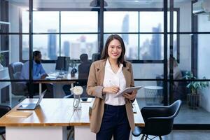 retrato do ásia o negócio CEO mulher é em pé dentro dela escritório às a mesa com digital tábua e mostrando estatística gráfico mostrando anual relatório e arranha-céu fundo foto