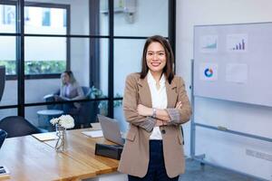 retrato do ásia o negócio CEO mulher é em pé mão cruzada dentro dela escritório às a mesa com estatística gráfico mostrando anual relatório foto