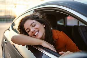 alegre jovem fêmea sentado dentro brilhante carro em passageiro assento e inclinado Fora aberto janela enquanto desfrutando a passeio foto
