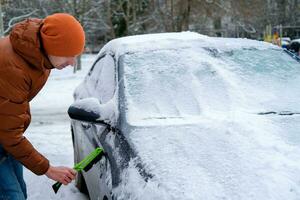 uma homem detém uma escova dentro dele mãos e limpa a janelas do a carro a partir de neve. a conceito do transporte, inverno, clima, pessoas e veículos. foto