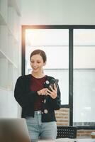 atraente jovem mulher falando em a inteligente telefone e sorridente enquanto em pé dentro escritório. foto