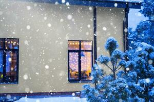 janela festiva aconchegante da casa do lado de fora com a luz quente das guirlandas de luzes de fada dentro - comemore o natal e o ano novo em um lar aconchegante. árvore de natal, bokeh, neve em pinheiros e queda de neve foto