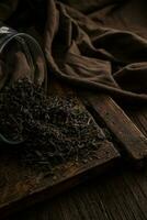 delicioso seco chá folhas espalhados a partir de uma jarra em uma de madeira vintage borda em uma Sombrio fundo, fechar-se foto