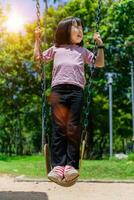 fofa pequeno menina tendo Diversão em uma balanço dentro lindo verão jardim. criança menina jogando em ao ar livre Parque infantil dentro parque. ativo verão lazer para crianças foto