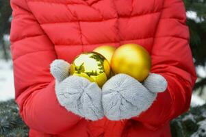 dourado bolas para a Natal árvore mentira dentro a mãos do uma mulher dentro caloroso cinzento luvas e uma vermelho jaqueta. festivo Novo ano decorações. foto