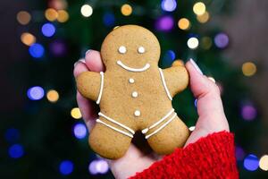 uma mulher mão detém uma Pão de gengibre homem em a fundo do a Natal árvore. faça você mesmo presentes. festivo humor. fechar-se. seletivo foco. foto