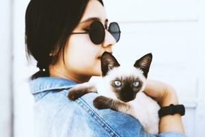 jovem atraente mulher abraçando bichano gato dentro mãos. fofa e glamouroso menina dentro na moda oculos de sol posando com dela siamês gato foto