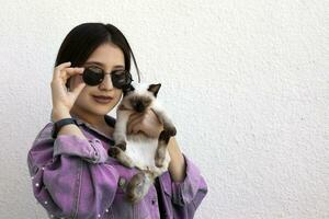 jovem atraente mulher abraçando bichano gato dentro mãos. fofa e glamouroso menina dentro na moda oculos de sol posando com dela siamês gato foto