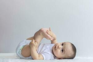 pequeno com fome bebê Garoto levando pés dentro boca. 6 meses bebê tendo Diversão dentro branco roupa de cama. fofa bebê deitado em cama. família, Novo vida, infância conceito. foto