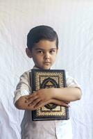 uma jovem meio Oriental Garoto com a piedosos alcorão. retrato do 5 anos velho muçulmano criança segurando uma piedosos Alcorão com branco fundo. livre espaço foto