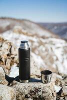 uma Preto garrafa térmica carrinhos em uma pedra dentro a montanhas. turista equipamento, equipamento para caminhada dentro natureza. inoxidável aço, chá caneca, montanhismo foto