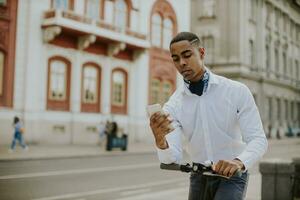 jovem afro-americano usando telefone celular em pé com uma scooter elétrica em uma rua foto