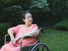 feliz ásia Senior mulher sentado em cadeira de rodas dentro a jardim, sorridente e olhando voltar. foto