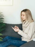 feliz jovem mulher relaxante em confortável sofá, segurando Smartphone dentro mãos. foto