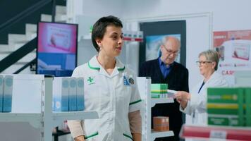 amigáveis farmacêutico dentro farmacêutico colocação pílulas caixa em prateleiras enquanto idosos colega de trabalho ajuda cliente com informações. sorridente profissional dentro farmácia arranjo medicinal produtos foto