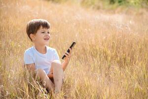 uma lindo criança sentado em a Relva fala de telefone dentro a verão às pôr do sol. Garoto comunica em Móvel foto