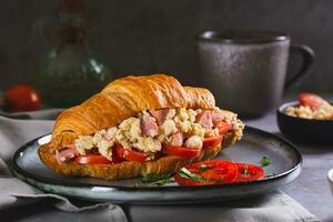 apetitoso croissant sanduíche com mexidos ovos, salsichas, tomates e ervas em uma prato foto