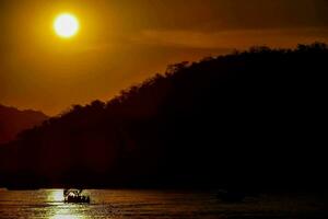 a Sol conjuntos sobre a água Como uma barco flutua dentro a distância foto