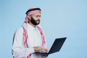 confuso muçulmano homem vestindo tradicional roupas trabalhando em computador portátil e digitando em teclado. árabe pessoa vestido dentro Thobe e lenço de cabeça segurando portátil computador e escrevendo mensagem foto