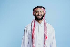 sorridente homem vestido dentro tradicional muçulmano roupas em pé com alegre expressão estúdio retrato. feliz árabe pessoa posando dentro Thobe e lenço de cabeça enquanto olhando às Câmera com despreocupado emoção foto