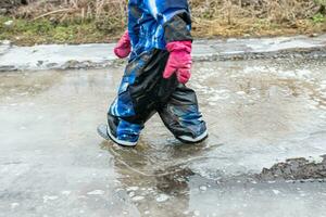 uma pequeno criança anda em através uma sujo poça dentro borracha chuteiras dentro a Primavera com peças do gelo foto