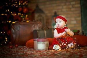 pequeno menina dentro vermelho vestir contra fundo do Natal árvore detém Natal festão dentro dela mãos. bebê 6 mês velho comemora Natal. foto