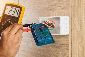 eletricista usando um medidor digital para medir a tensão em uma tomada em uma parede de madeira.