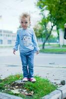 pequeno lindo menina com curto encaracolado cabelo em pé em uma toco dentro a cidade. criança dentro jeans e uma azul camisa jogando ao ar livre foto