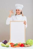 alegre fêmea chefe de cozinha é mostrando Está bem placa enquanto segurando quadro branco com grupo do legumes em a mesa. foto