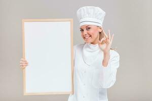 retrato do lindo fêmea chefe de cozinha mostrando Está bem placa enquanto segurando quadro branco em cinzento fundo. foto