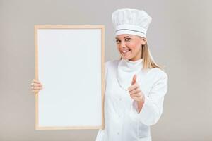retrato do lindo fêmea chefe de cozinha mostrando polegar acima enquanto segurando quadro branco em cinzento fundo. foto