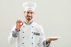 sorridente chefe de cozinha é segurando prato e mostrando Está bem placa em cinzento fundo foto