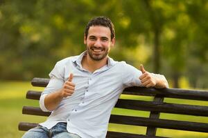 jovem homem de negocios é sentado às a parque e mostrando polegar acima foto
