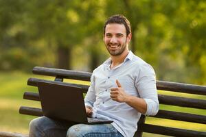 jovem homem de negocios é sentado às a parque e usando computador portátil e mostrando polegar acima foto