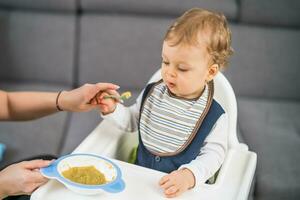 fofa bebê Garoto Aprendendo para comer de ele mesmo enquanto sentado dentro uma Alto cadeira foto