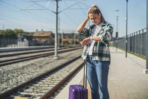 chateado mulher olhando às dela relógio enquanto em pé com mala de viagem em uma trem estação foto