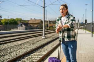 chateado mulher olhando às dela relógio enquanto em pé com mala de viagem em uma trem estação foto
