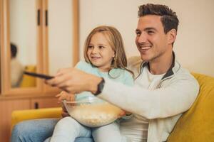 pai e filha apreciar assistindo televisão e comendo pop calos juntos às seus casa foto
