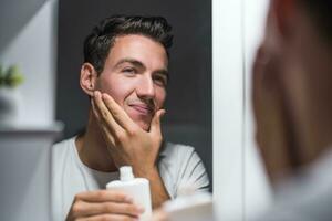 homem é aplicando loção pós-barba enquanto olhando ele mesmo dentro a espelho foto