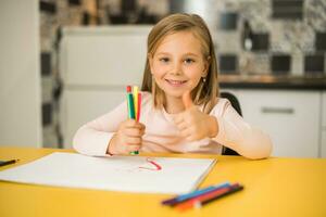 lindo pequeno menina mostrando polegar acima enquanto goza desenhando às dela casa foto