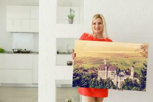 uma feliz jovem Loiras mulher é segurando uma ampla parede foto tela de pintura às casa