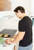 adulto homem em pé perto cozinhando alcance e corte tomate dentro cozinha foto