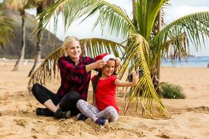 mãe e filha dentro santa chapéu levar selfie em a de praia perto Palma árvore foto