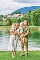 ao ar livre retrato do lindo família, jovem casal com pré-escolar Garoto e criança pequena menina posando Próximo para lago ou rio foto