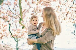 ao ar livre retrato do feliz jovem mãe com adorável bebê menina debaixo florescendo Primavera árvore, inferior Visão foto