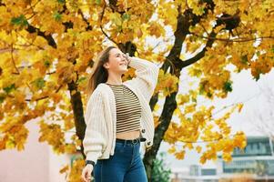 outono retrato do jovem bonita menina posando Próximo para árvore com amarelo folhas, feliz humor e saudável estilo de vida foto