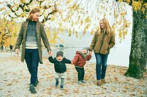 ao ar livre retrato do jovem feliz família do quatro, mãe e pai jogando com crianças dentro outono parque de a lago, frio clima foto