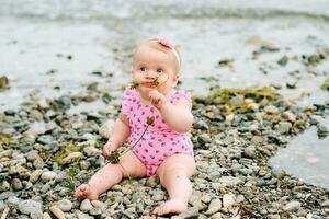 ao ar livre retrato do adorável bebê menina jogando com algas marinhas de a rio, vestindo Rosa roupa de banho foto