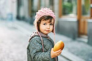 ao ar livre retrato do fofa criança pequena menina segurando leite pão pão, vestindo caloroso Jaqueta e chapéu, lanche para criança foto