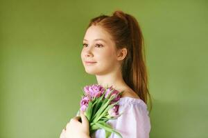 beleza retrato do bonita jovem 15 - 16 ano velho ruivo adolescência menina vestindo roxa vestir posando em verde fundo, segurando tulipa flores foto